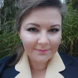 Portret kobiety z zielonymi kolczykami na tle zieleni, ubrana w granatową marynarkę z beżowym kołnierzem.