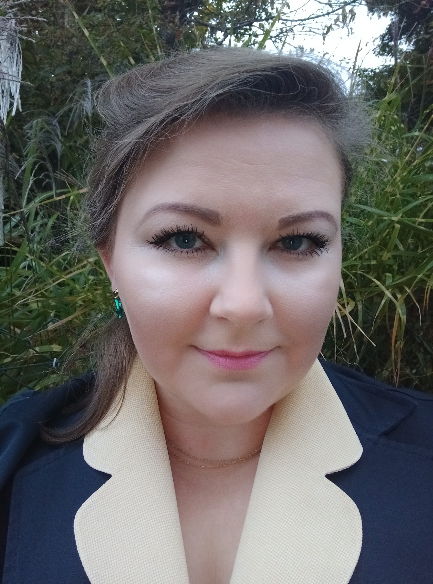Portret kobiety z zielonymi kolczykami na tle zieleni, ubrana w granatową marynarkę z beżowym kołnierzem.