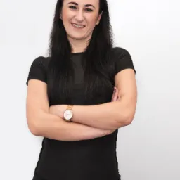 Uśmiechnięta brunetka w czarnym sportowym stroju, ze skrzyżowanymi ramionami i złotym zegarkiem na nadgarstku, pozuje na jasnym tle.