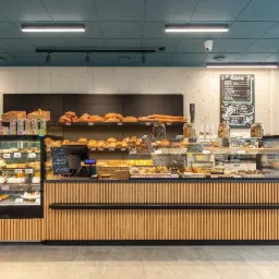 Wnętrze piekarni-cukierni: lady chłodnicze z ciastami i tortami, półki z pieczywem, tablica z menu kawy i herbaty, drewniane wykończenie.