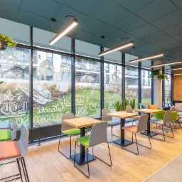 Nowoczesna przestrzeń piekarnio-cukierni z drewnianymi stolikami i krzesłami w pastelowych kolorach, duże okna z dekoracyjnymi napisami i grafikami, wiszące lampy liniowe i rośliny doniczkowe.