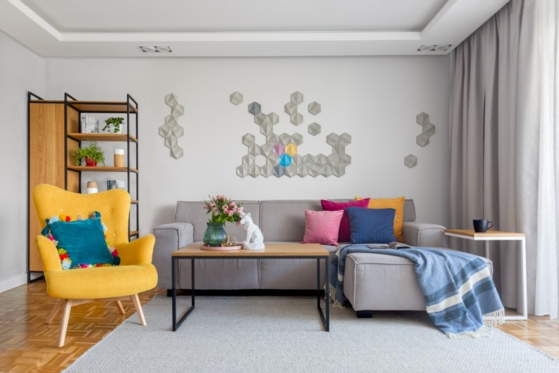 Jasne wnętrze salonu z szarą sofą, żółtym fotelem z turkusową poduszką, drewnianym regałem i dekoracją ścienną z heksagonów w odcieniach szarości z akcentami kolorystycznymi.