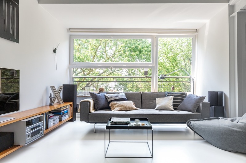 Jasne wnętrze apartamentu z szarą sofą, stolikiem kawowym, regałem RTV i dużym oknem z widokiem na zieleń. Na stoliku filiżanka i książki. Minimalistyczny design.