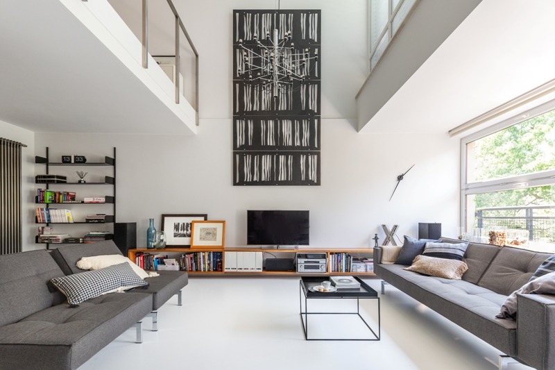 Dwupoziomowy apartament w jasnej tonacji z szarą sofą, drewnianą szafką RTV i wysokim oknem z widokiem na zieleń, uzupełniony nowoczesną grafiką na ścianie.