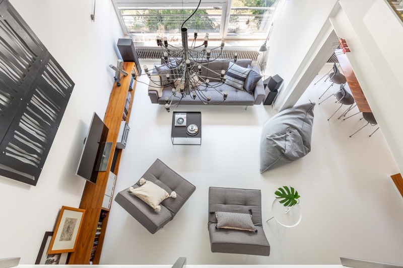 Dwupoziomowy apartament widziany z góry: szara sofa z poduszkami, designerski żyrandol, dwa fotele z poduszkami, pufa w kształcie worka i długa drewniana półka z telewizorem.