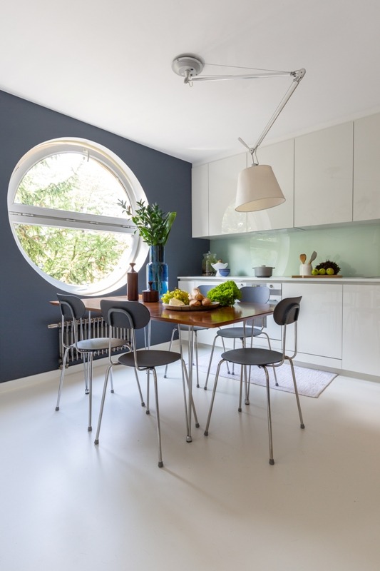 Dwupoziomowy apartament na Sadybie: stół jadalny z owocami i warzywami pod wiszącą lampą, okrągłe okno z widokiem na zieleń, grafitowa ściana.