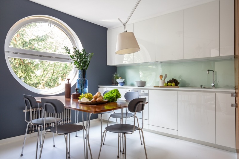 Dwupoziomowy apartament w Warszawie Sadybie: drewniany stół z przekąskami i bukietem, okrągłe okno z widokiem na zieleń, biała zabudowa kuchenna z zielonym panelem, nowoczesne krzesła i lampa.