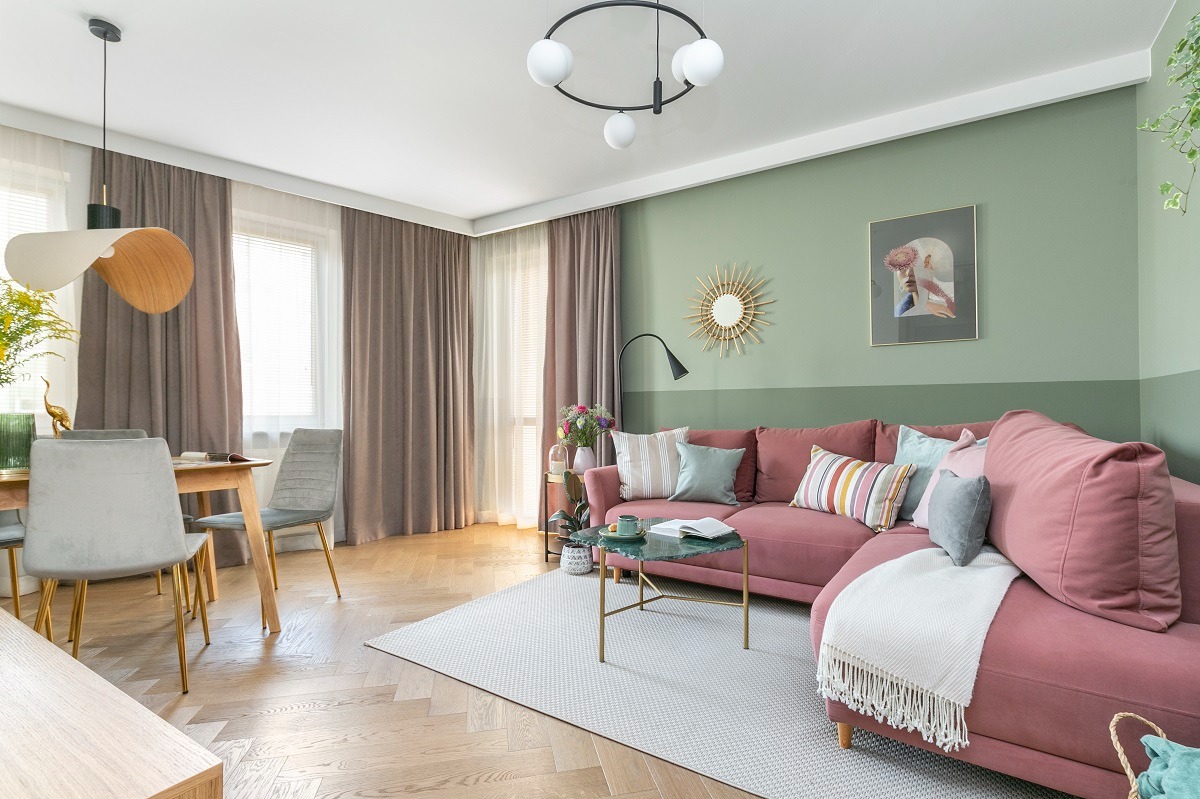 Jasne mieszkanie w Krakowie: różowa sofa narożna z poduszkami, stolik kawowy z marmurowym blatem, dywan w geometryczny wzór, drewniana podłoga w jodełkę, stół z krzesłami oraz designerskie lampy...