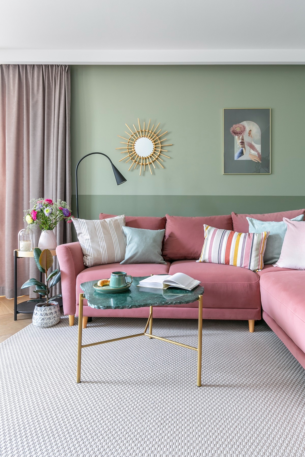 Salon w odcieniach różu i zieleni: narożna sofa, stolik z zielonym blatem, dywan w geometryczny wzór, ściana dwukolorowa z lustrem-słońcem i obrazem.