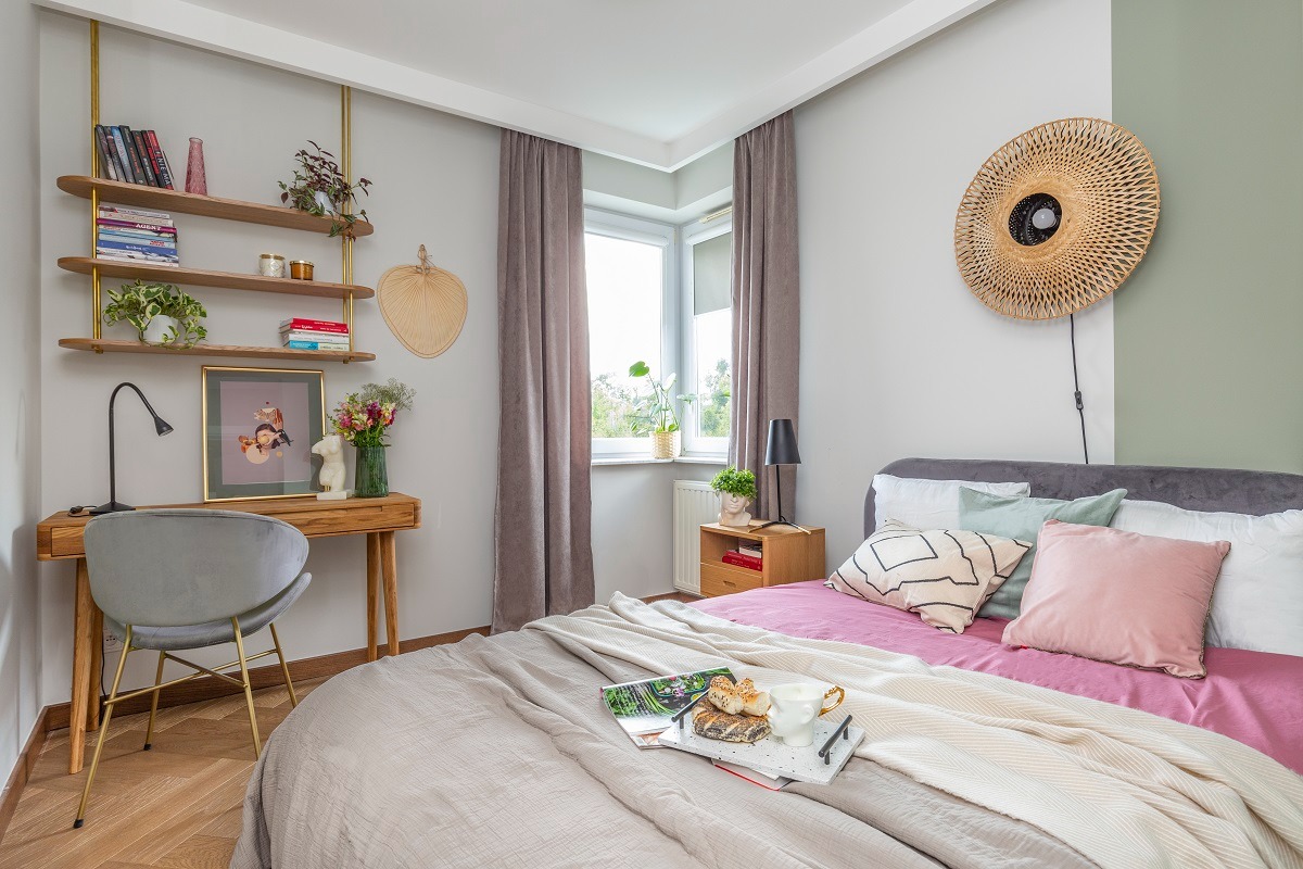 Jasna sypialnia z drewnianym biurkiem i półkami, szarym krzesłem na złotych nogach, łóżkiem z różową pościelą i dekoracyjnym, okrągłym abażurem z rattanu na ścianie.