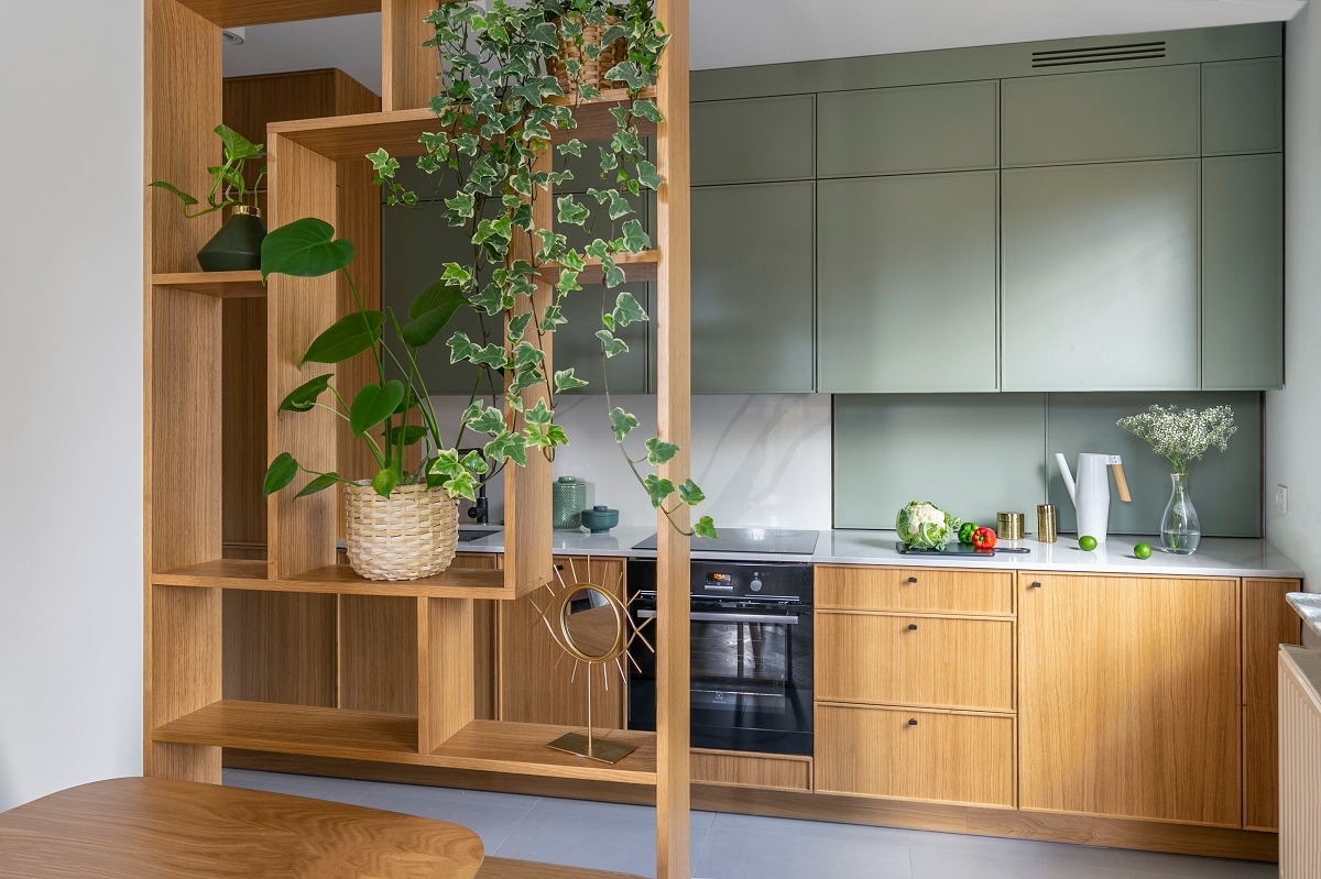 Nowoczesna kuchnia z drewnianą zabudową, zielonymi szafkami i roślinami doniczkowymi na ażurowym regale, dzielącym przestrzeń. Na blacie biały dzbanek i warzywa.