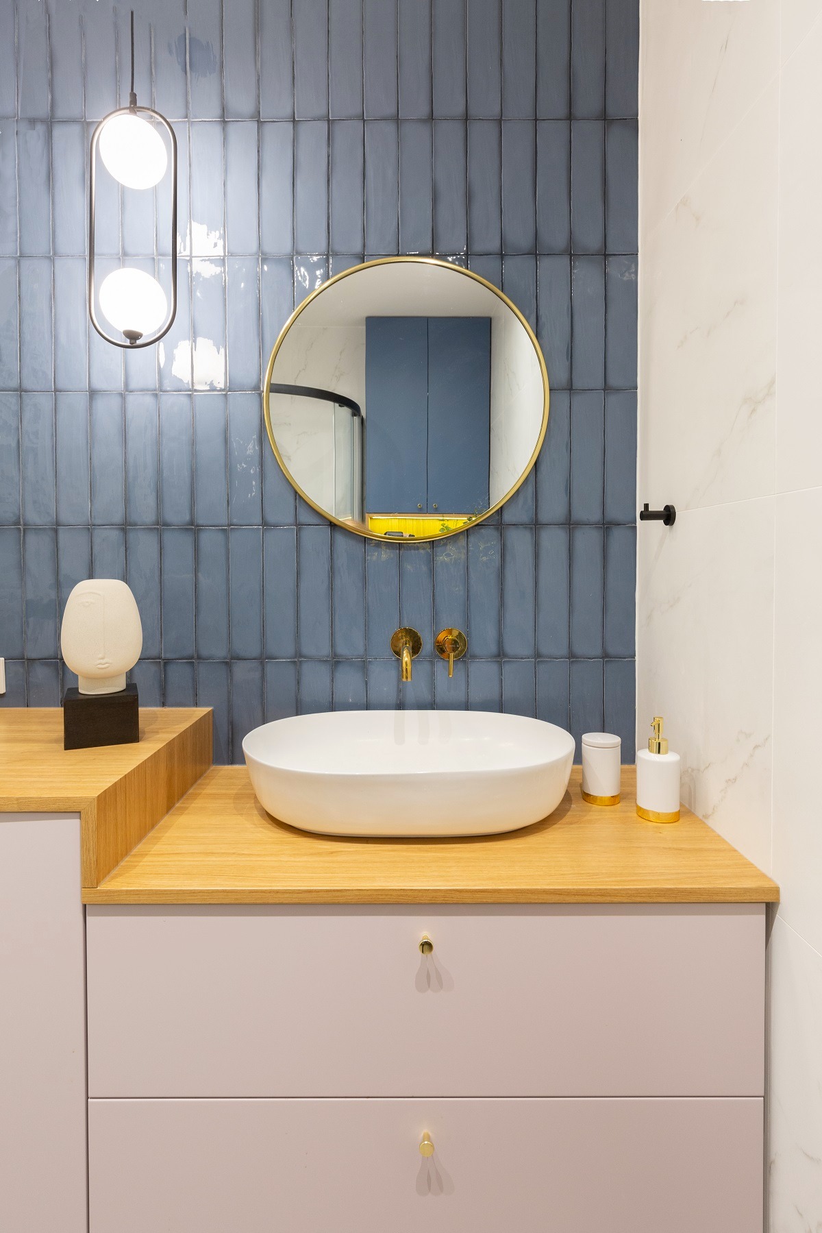 Nowoczesna łazienka z owalną umywalką nablatową, złotą armaturą, niebieskimi płytkami wertykalnymi i okrągłym lustrem ze złotą ramą, uzupełniona o dekoracyjną rzeźbę.