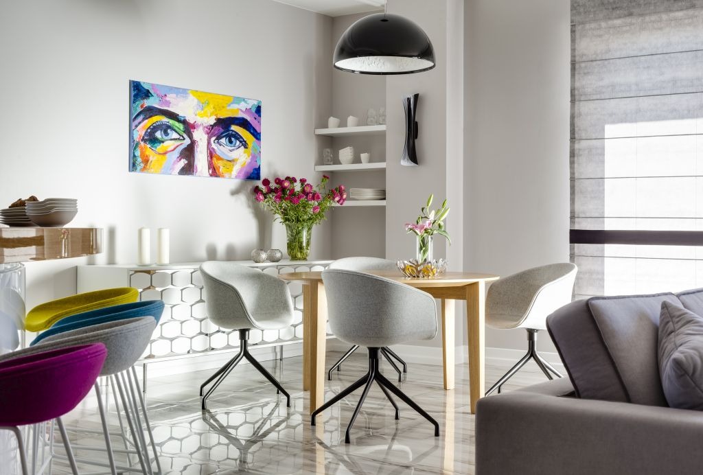 Jasna jadalnia w apartamencie: drewniany stół otoczony szarymi krzesłami na czarnych nogach, nad stołem czarna lampa, na ścianie abstrakcyjny obraz z motywem oczu, w tle biały kredens i półki...