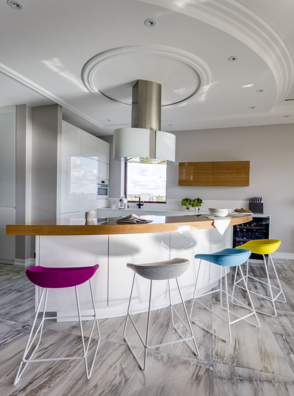 Nowoczesna kuchnia z wyspą, drewnianym blatem i kolorowymi hokerami. Widoczna okrągła dekoracja sufitowa z oświetleniem i stalowym okapem.