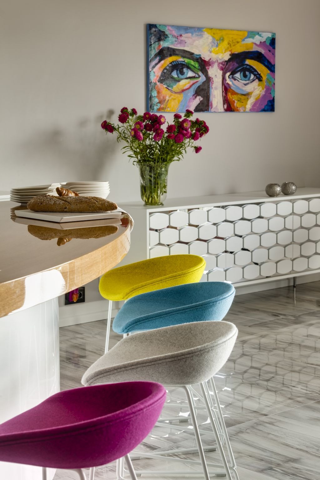 Nowoczesna kuchnia z czterema kolorowymi krzesłami (fioletowy, szary, niebieski, żółty) ustawionymi przy blacie, na blacie chleb i talerze, w tle biała komoda z geometrycznym wzorem i obraz...