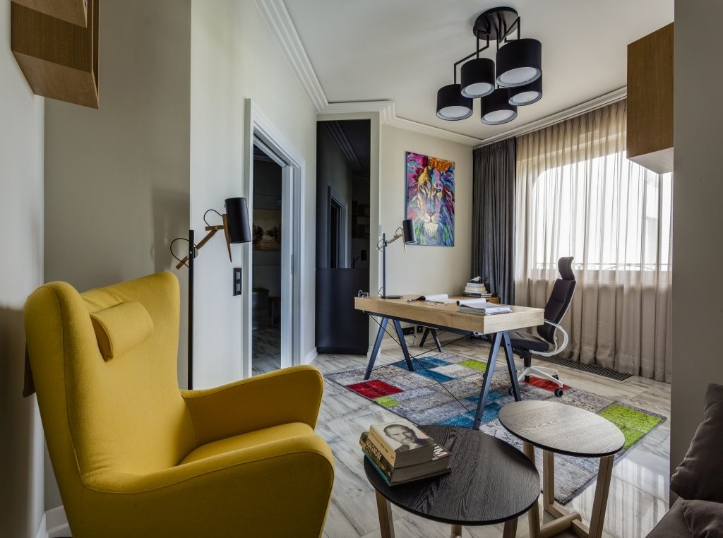 Nowoczesny gabinet w apartamencie: żółty fotel, biurko z czarnymi nogami, kolorowy dywan patchwork, grafika lwa na ścianie, marmurowa podłoga i czarne stoliki kawowe.