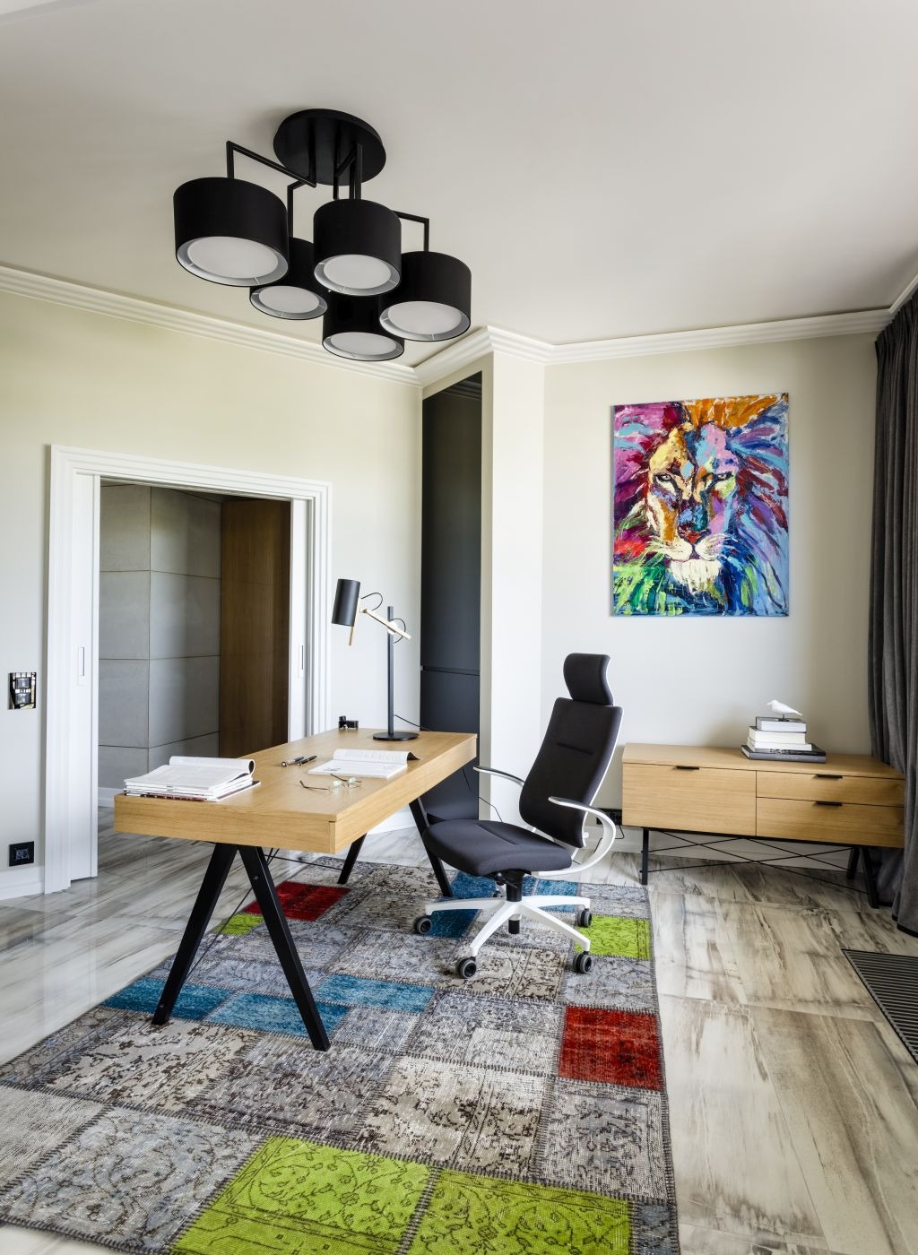 Jasne biuro domowe z drewnianym biurkiem na czarnych nogach, krzesłem biurowym na kółkach, wzorzystym dywanem i obrazem lwa na ścianie.