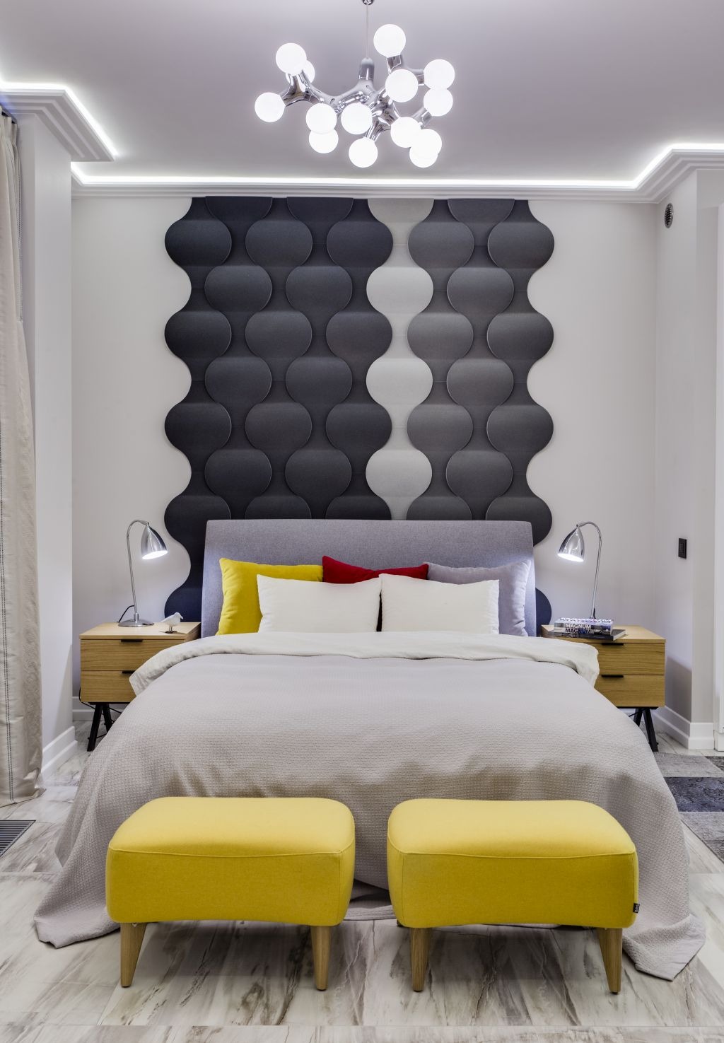 Nowoczesna sypialnia z szarym łóżkiem, żółtymi pufami i geometrycznym panelem ściennym w odcieniach szarości. Lampki nocne na drewnianych szafkach, modernistyczny żyrandol.