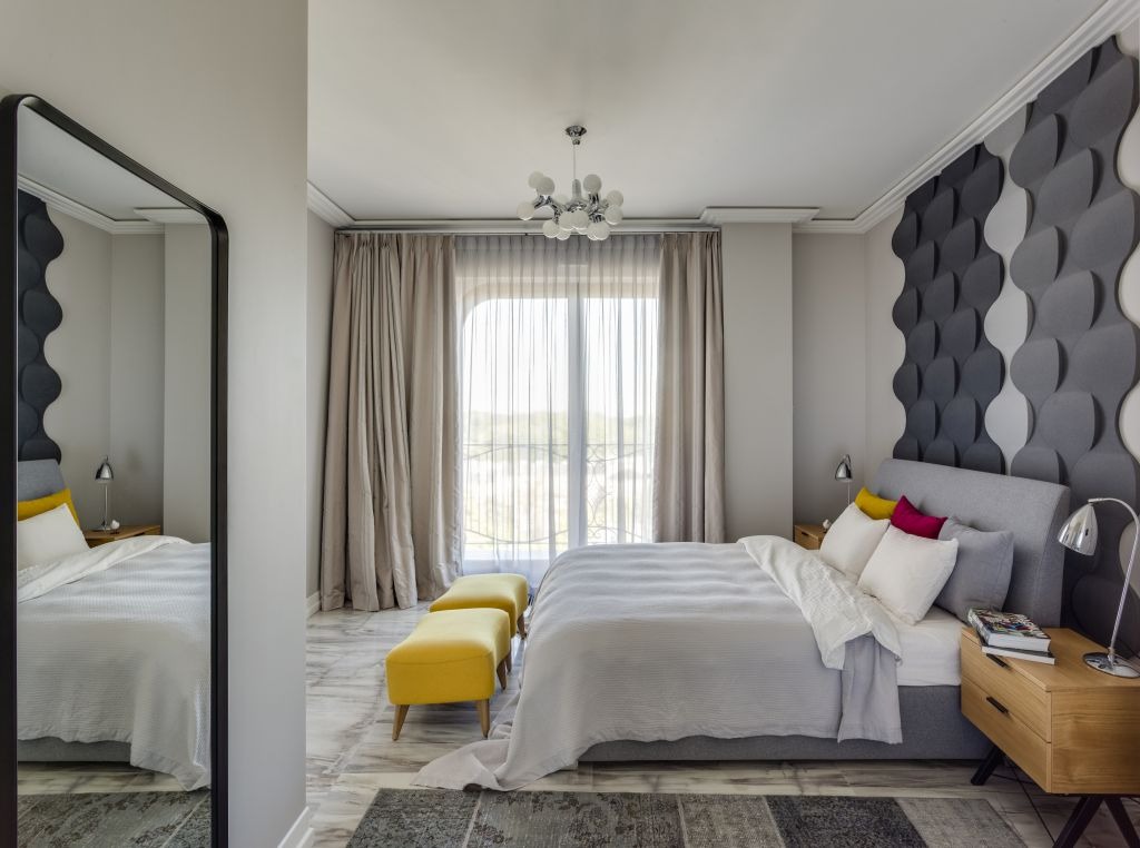 Nowoczesna sypialnia z łóżkiem, szarą tapetą 3D, żółtymi pufami i dużym lustrem odbijającym wnętrze, widok na balkon przez okno z beżowymi zasłonami.