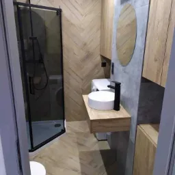 Nowoczesna łazienka z kabiną prysznicową typu walk-in, umywalką nablatową na drewnianym blacie, lustrem owalnym i płytkami ułożonymi w jodełkę na ścianie. Widoczna pralka w tle.