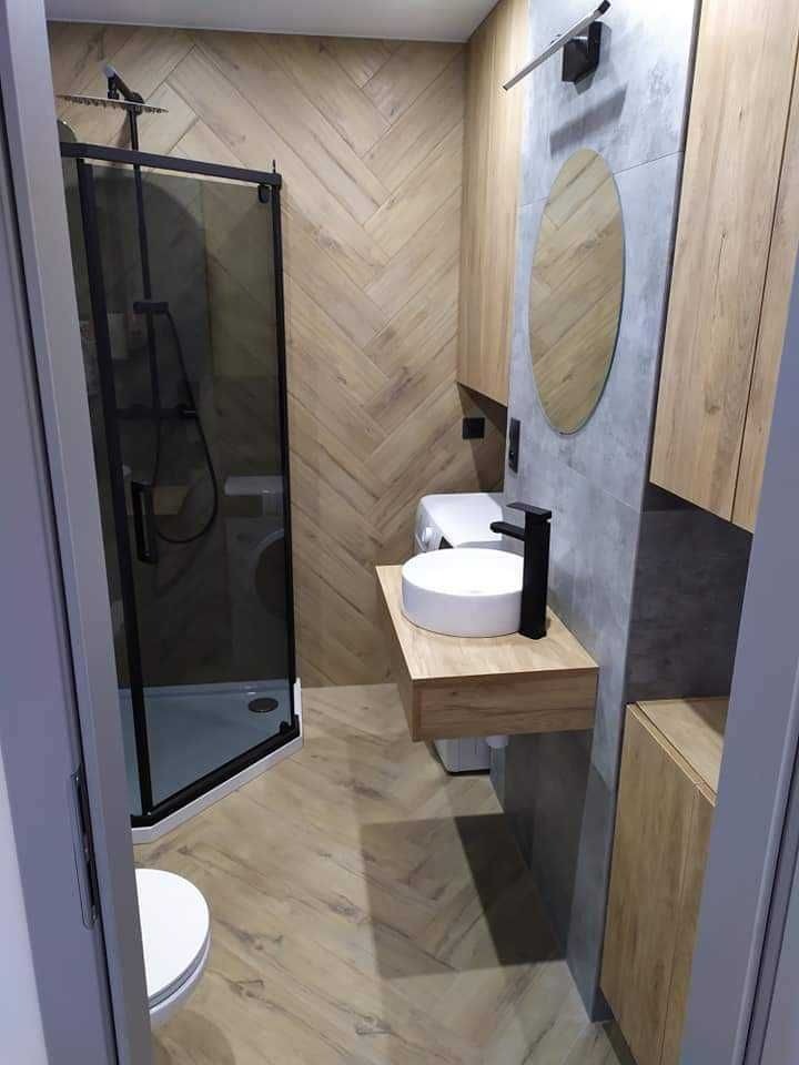 Nowoczesna łazienka z kabiną prysznicową typu walk-in, umywalką nablatową na drewnianym blacie, lustrem owalnym i płytkami ułożonymi w jodełkę na ścianie. Widoczna pralka w tle.