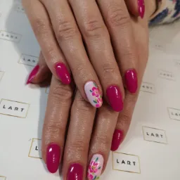 Dłonie z owalnymi paznokciami pomalowanymi na intensywny różowy kolor, jeden paznokieć na każdej dłoni zdobiony motywem kwiatowym na jasnoróżowym tle, na tle papieru z powtarzającym się logo.