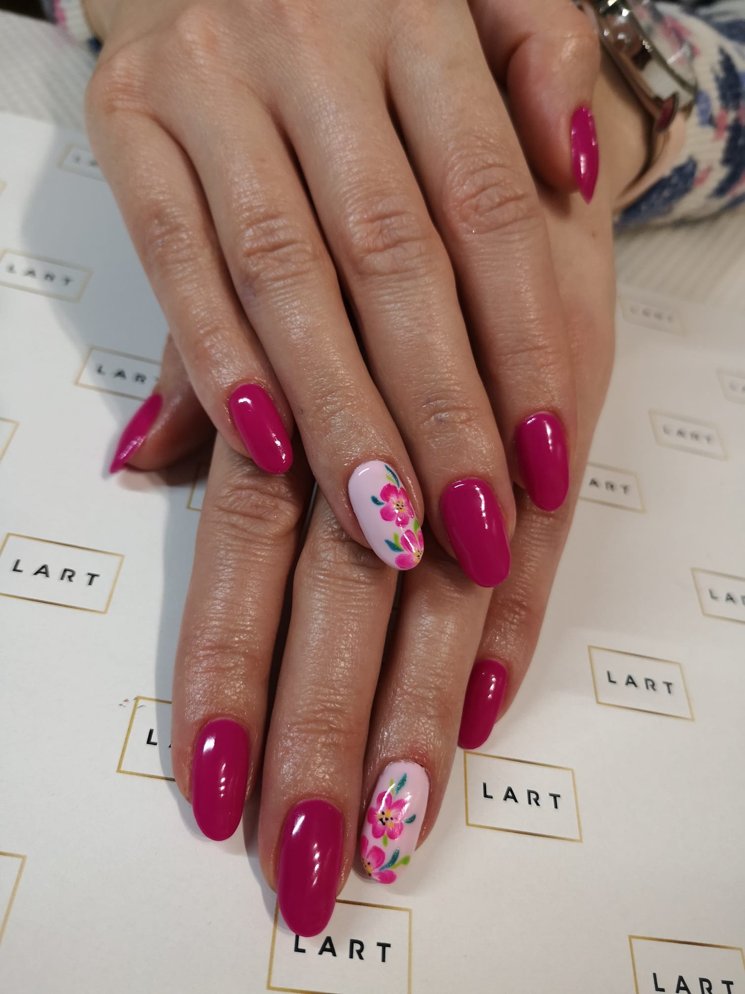 Dłonie z owalnymi paznokciami pomalowanymi na intensywny różowy kolor, jeden paznokieć na każdej dłoni zdobiony motywem kwiatowym na jasnoróżowym tle, na tle papieru z powtarzającym się logo.