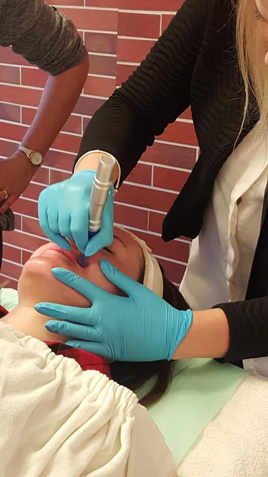 Zabieg na twarzy z użyciem urządzenia w gabinecie kosmetycznym, widoczne turkusowe rękawiczki ochronne i fragment ściany z cegieł.