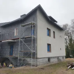 Dwukondygnacyjny dom w trakcie ocieplania elewacji, część ściany pokryta styropianem, a część otynkowana, widoczne rusztowanie i pracownik w górnej części rusztowania. Okna zabezpieczone folią.