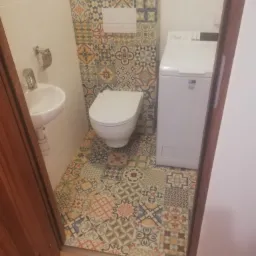 Ukończona łazienka z wzorzystymi płytkami podłogowymi i ściennymi, białą umywalką narożną, toaletą i pralką, widok zza otwartych drzwi.