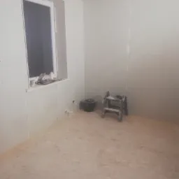 Pokój w trakcie prac wykończeniowych: ściany obłożone płytami kartonowo-gipsowymi, widoczne łączenia, okno z prowizorycznym parapetem, drabina malarska i wiadro na podłodze.