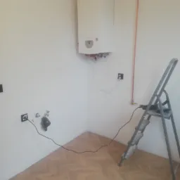 Biały piec gazowy zamontowany na białej ścianie w rogu pokoju z parkietem; widoczne miedziane rury, gniazdka elektryczne, podłączone przewody i aluminiowa drabina.