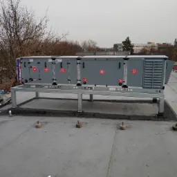 Centrala wentylacyjna na dachu budynku, widoczne elementy sterujące i oznaczenia, stalowa konstrukcja wsporcza, w tle drzewa i budynki miejskie.