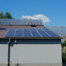 Panele słoneczne na dachu budynku z oranżerią, widoczne również rynna, czujnik alarmowy i kwietnik z kolorowymi kwiatami.