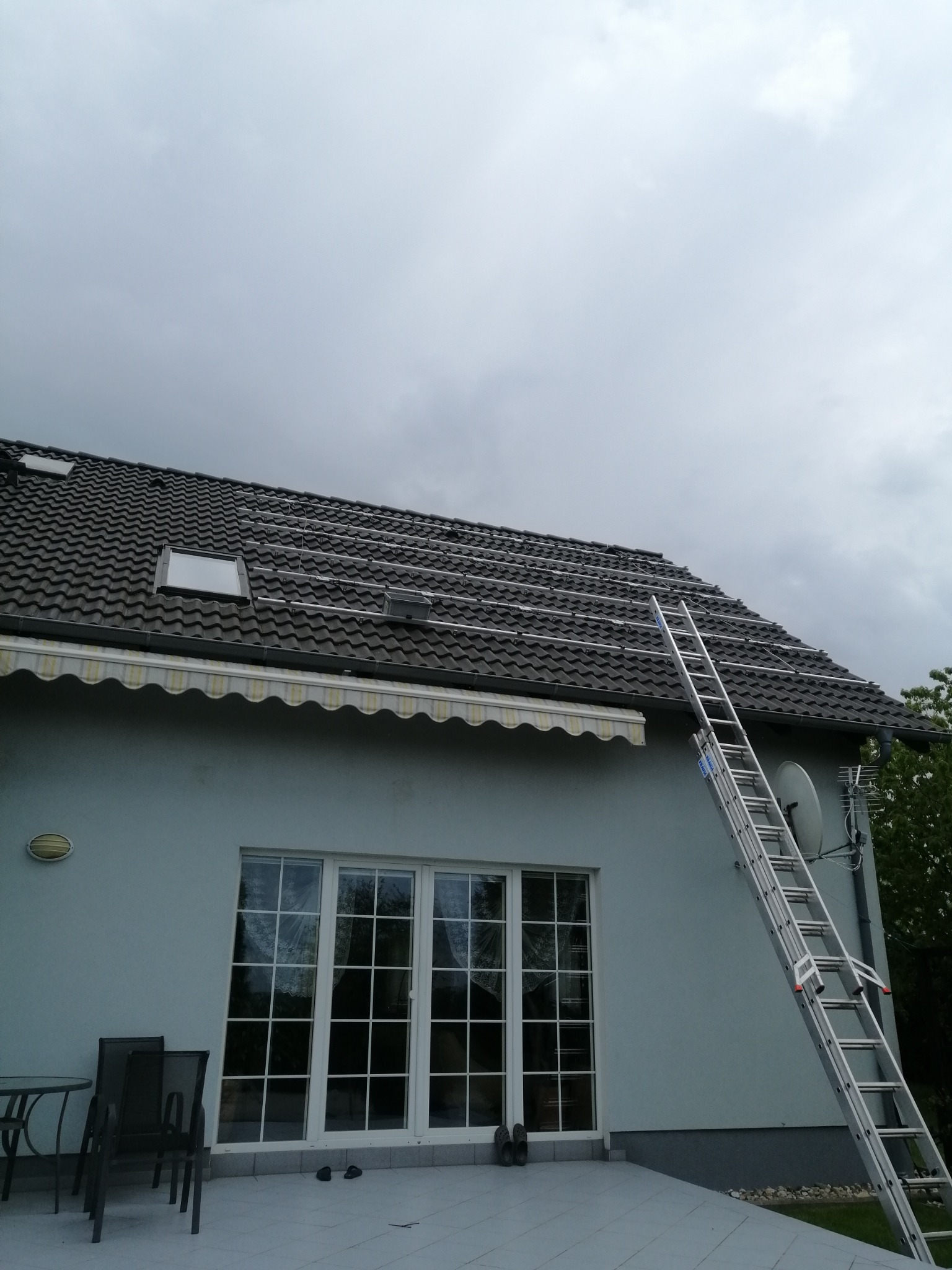 Montaż szyn pod panele słoneczne na dachu domu z ciemną dachówką, drabina oparta o ścianę, pochmurne niebo.