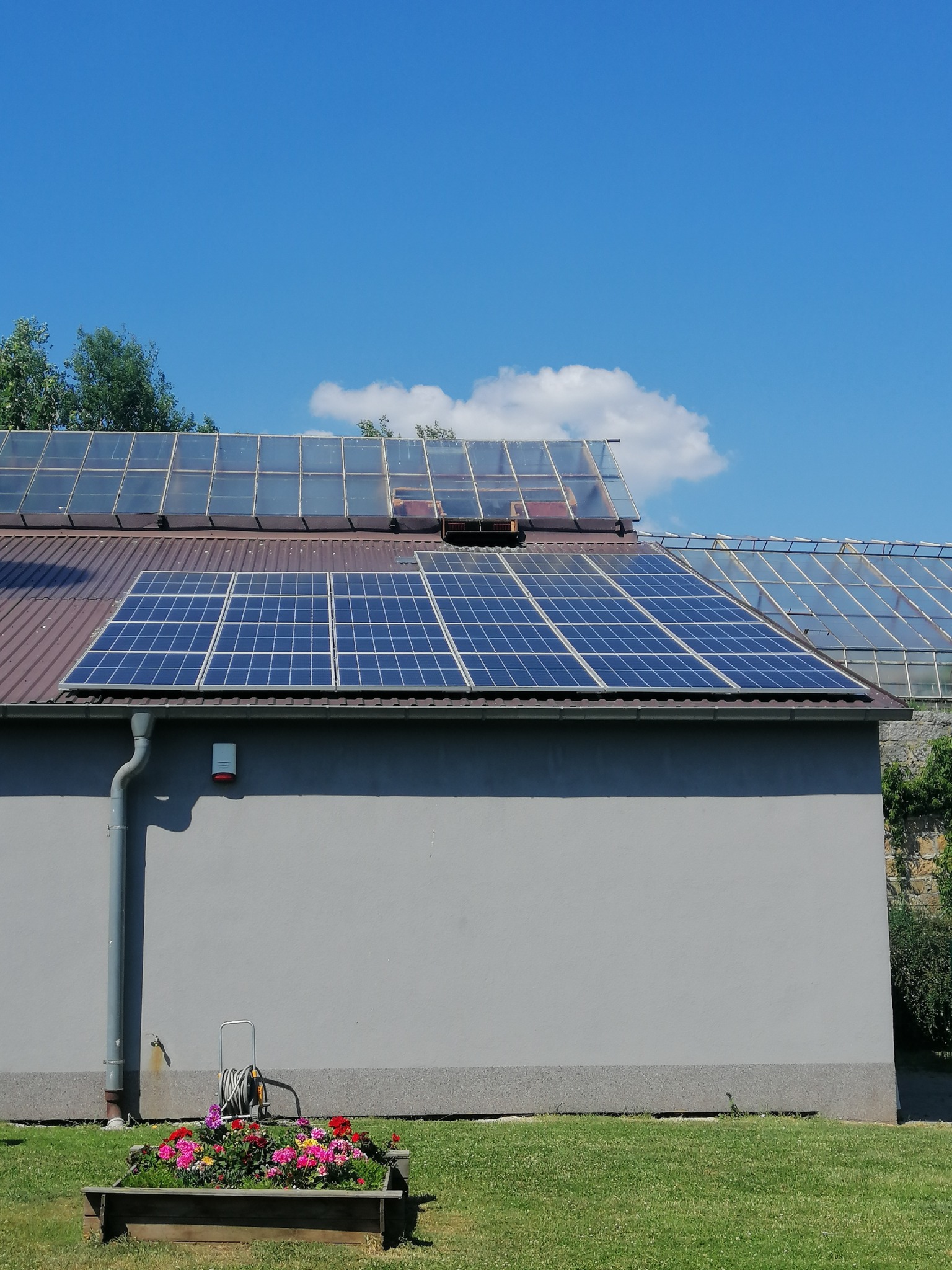 Panele słoneczne na dachu budynku z oranżerią, widoczne również rynna, czujnik alarmowy i kwietnik z kolorowymi kwiatami.