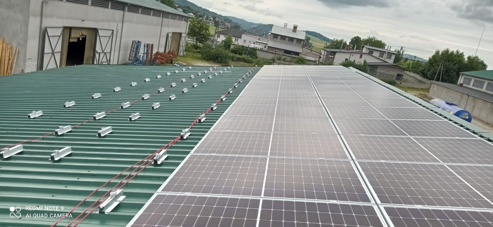 Montaż paneli fotowoltaicznych na zielonym dachu z blachy trapezowej, widoczne kable i elementy mocujące, w tle budynki gospodarcze i krajobraz wiejski.