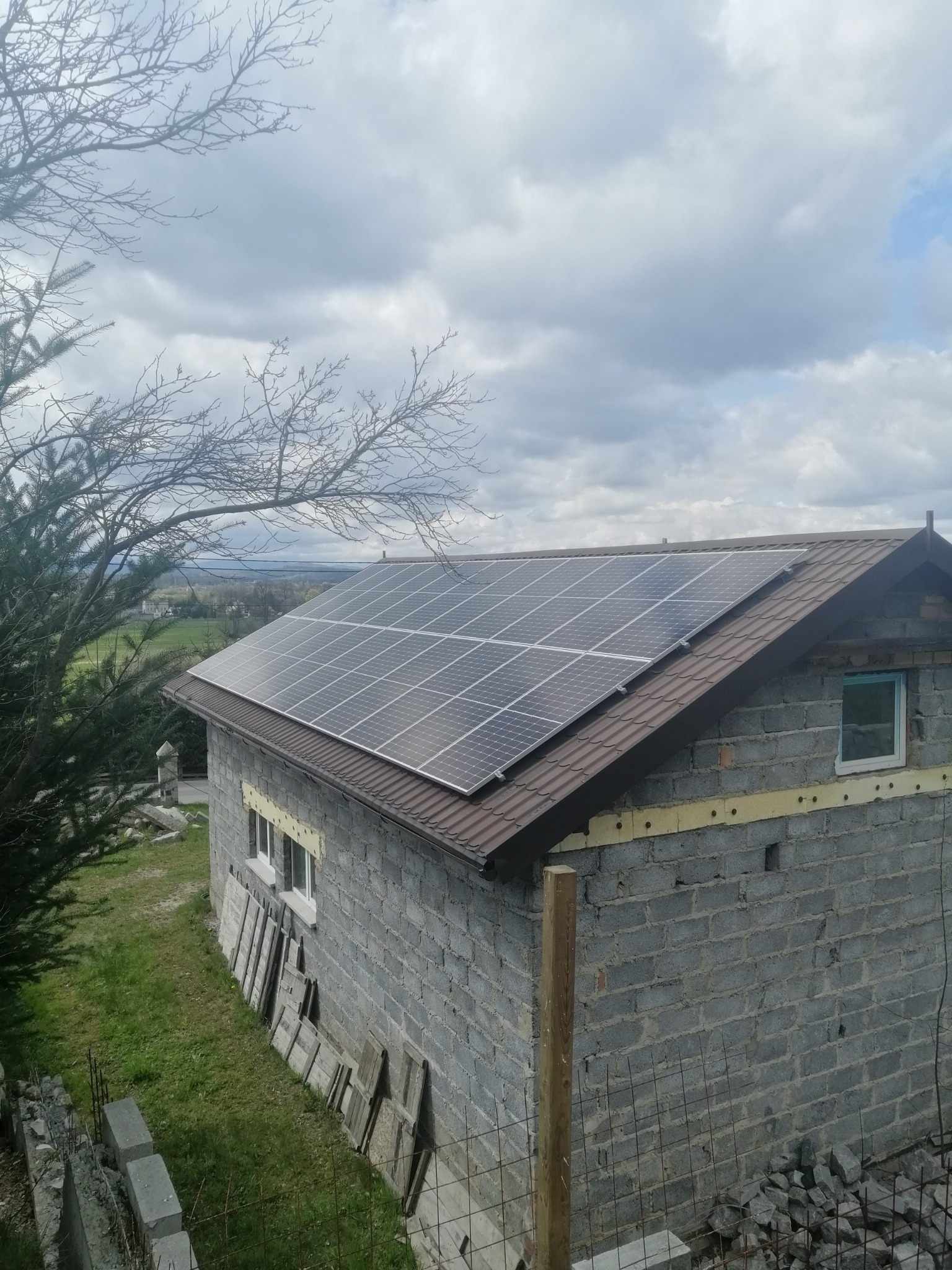 Dom w trakcie budowy z zamontowanymi panelami słonecznymi na brązowym dachu, widok z góry, pochmurne niebo w tle.