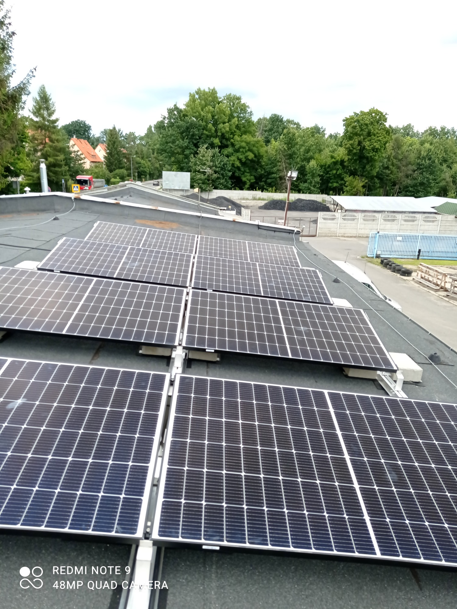 Rząd paneli słonecznych na płaskim dachu budynku przemysłowego, widok z góry, w tle drzewa i droga.