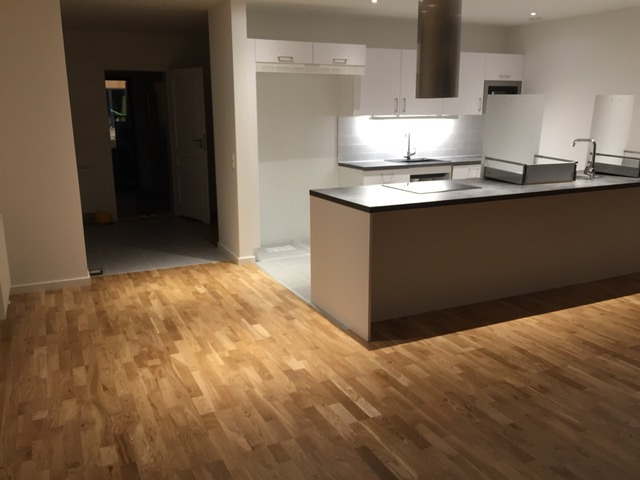 Jasny, dębowy parkiet ułożony w jodełkę francuską, widoczny na tle nowoczesnej kuchni z wyspą i białymi szafkami, z otwartym przejściem do innego pomieszczenia z szarą podłogą.