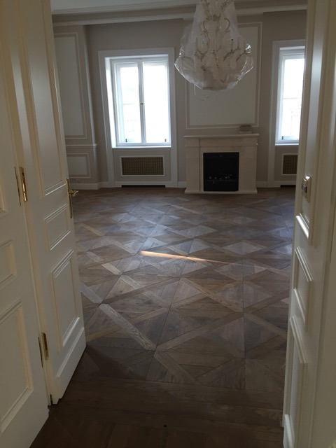 Wzorzysta drewniana podłoga w jodełkę, widziana zza uchylonych białych drzwi, z fragmentem żyrandola w tle.