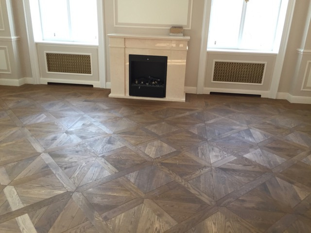 Parkiet ułożony we wzór geometryczny, z widocznymi detalami słojów drewna i marmurowym kominkiem z elektrycznym wkładem w tle.