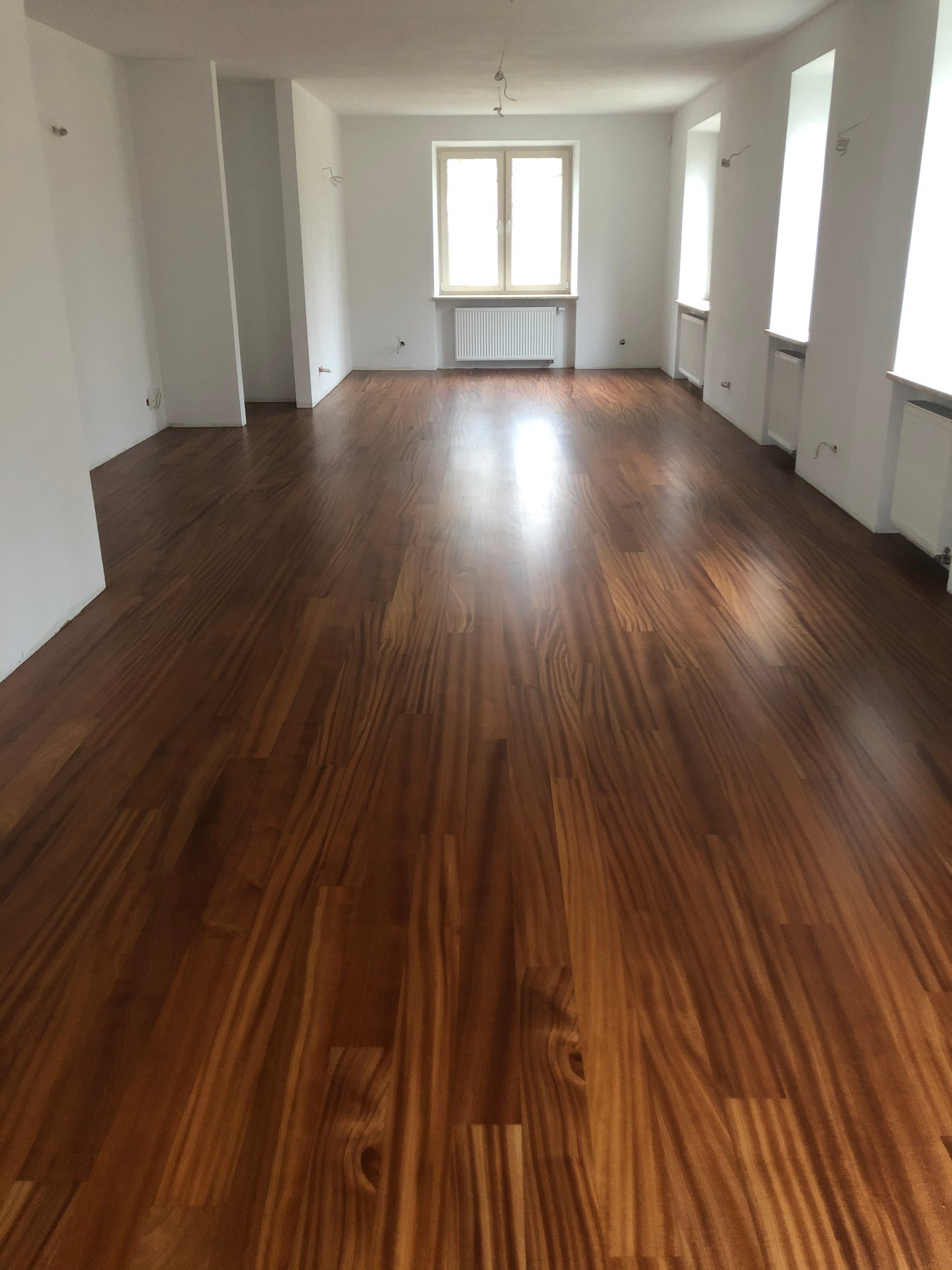 Wykończona podłoga z drewna egzotycznego w pustym, odnowionym pokoju w Warszawie, z widocznymi grzejnikami pod oknami.