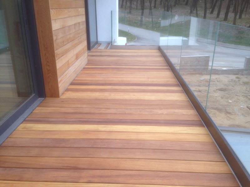 Drewniany taras z desek o zróżnicowanym odcieniu brązu, zabezpieczony szklaną balustradą, przylegający do nowoczesnego budynku z drewnianą elewacją i dużym oknem.