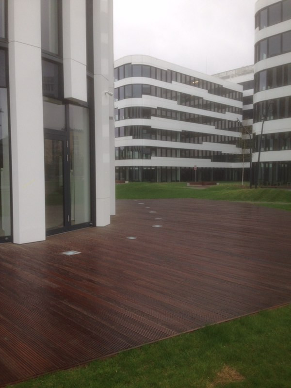 Ciemnobrązowy taras z desek kompozytowych na zewnątrz nowoczesnego biurowca z trawnikiem, z wbudowanymi punktowymi lampami podłogowymi.
