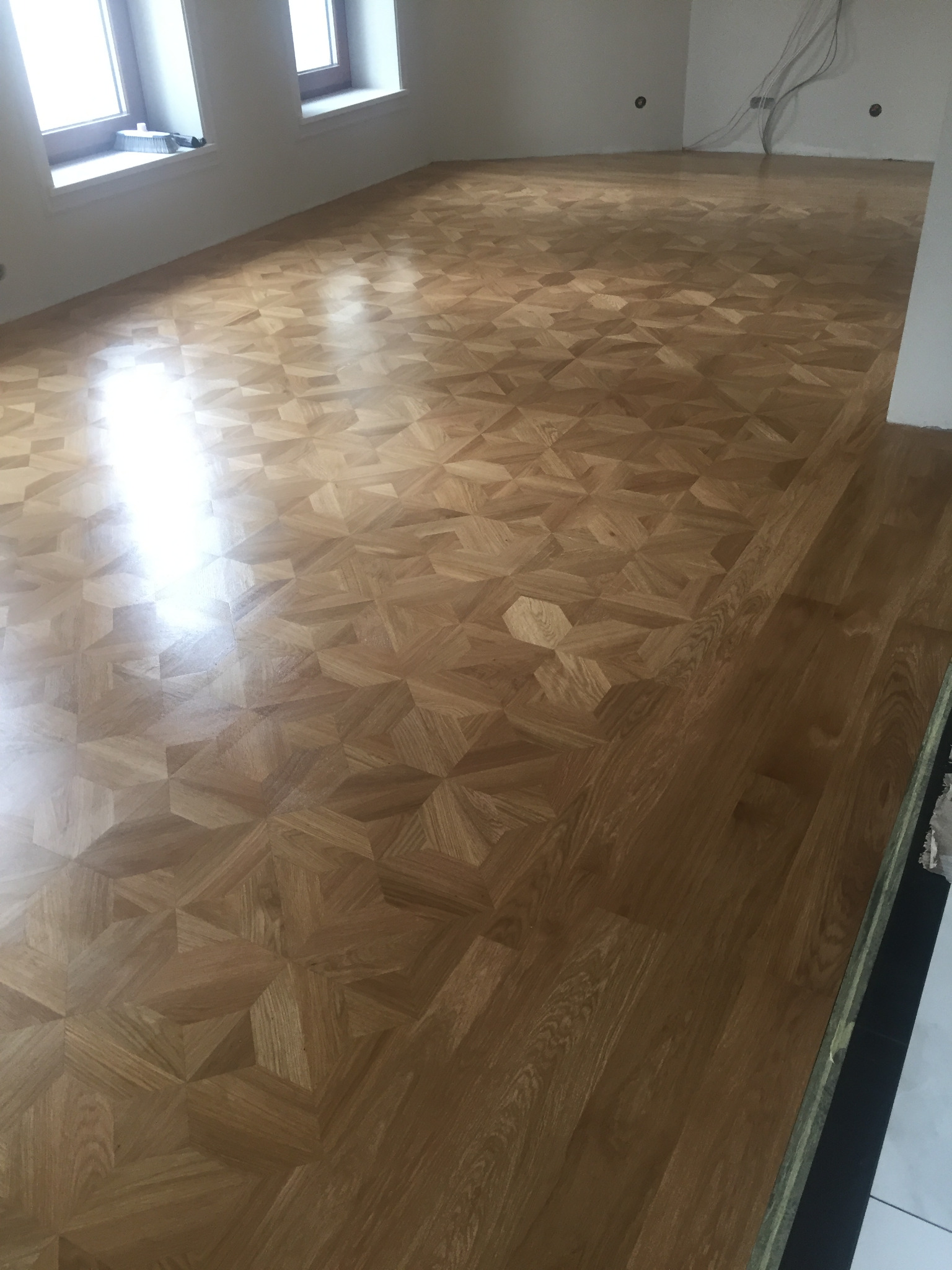 Jasny parkiet ułożony we wzór gwiazdy w przestronnym, słonecznym pokoju z białymi ścianami i dwoma oknami.