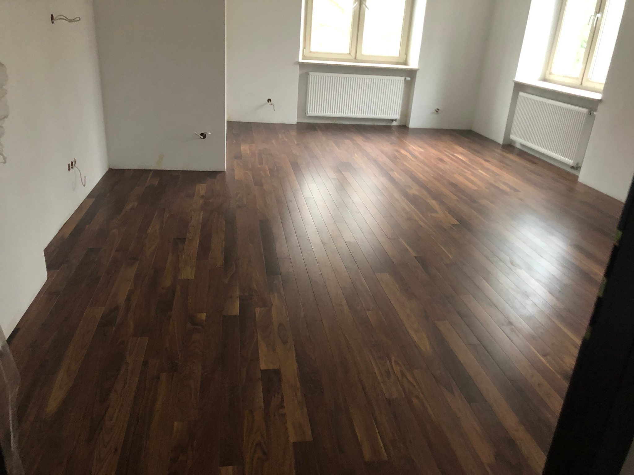 Ułożony parkiet z ciemnego drewna w jasnym pomieszczeniu z grzejnikami pod oknami, widoczne wystające kable ze ściany.