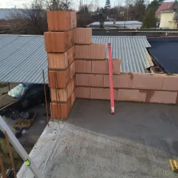 Wznoszenie ściany z czerwonych pustaków ceramicznych na betonowym stropie, widoczny poziomica w pionie, zbrojenie i fragment dachu z blachy falistej w tle.