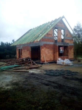 Wznoszenie ścian parterowego domu z bloczków ceramicznych, widoczna konstrukcja więźby dachowej z łatami, przygotowane materiały budowlane na placu budowy.
