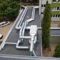 Widok z góry na system srebrnych kanałów wentylacyjnych na dachu budynku, z przyległym terenem zielonym i budynkami w tle.